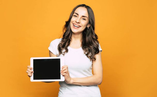 Here it is! A girl with sincere brown eyes, dark-brown wavy long hair near the vibrant orange background is holding a tablet PC in her elegant hands. - Fotografie, Obrázek