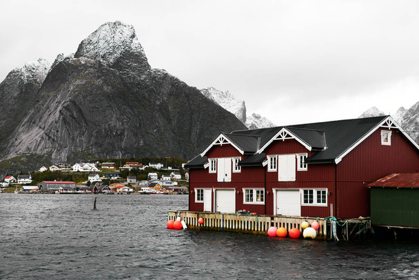 Traditional red wooden house at the coast in Reine harbor in front of a snowcapped mountain and another village. Reine is located in Moskenesoya on Lofoten Islands in Norway. - Photo, Image