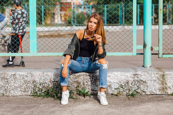 Fashion portrait of trendy young woman wearing sunglasses, and bomber jacket sitting next to rabitz in the city - Photo, Image