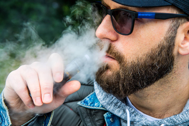 man with a beard smokes an electronic cigarette in a park - Photo, Image