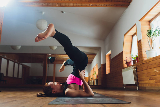 Young sporty attractive woman practicing yoga, doing Salamba Sarvangasana exercise, supported Shoulder stand pose, working out, wearing sportswear, pants and top, indoor close up, white yoga studio. - 写真・画像