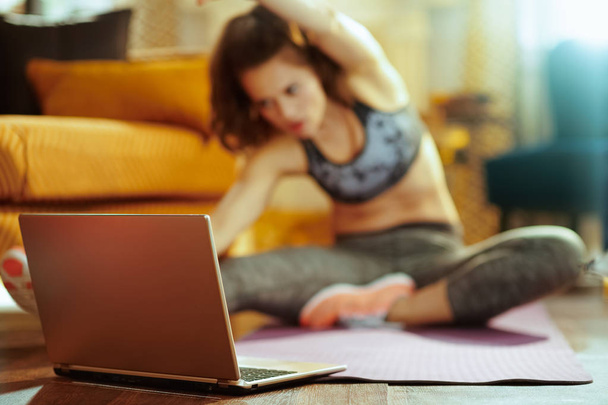 Gros plan sur ordinateur portable beige et femme en arrière-plan étirant sur tapis de fitness tout en regardant tutoriel de fitness sur Internet dans la maison moderne
. - Photo, image
