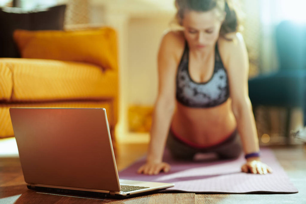 Closeup on beige laptop and woman in background stretching on fitness mat while watching fitness videos on internet in the modern house. - Φωτογραφία, εικόνα