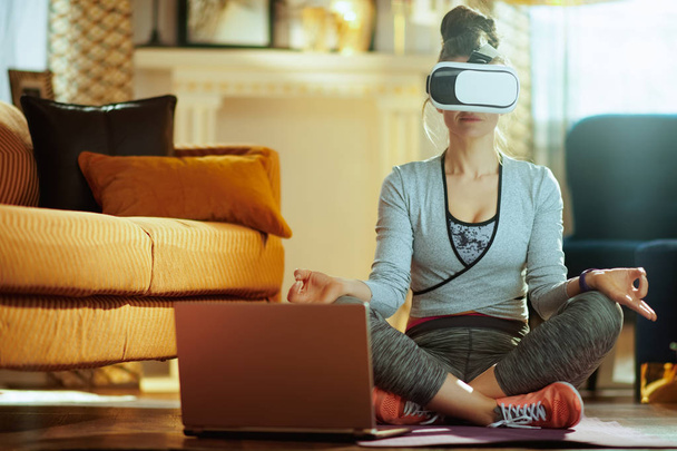 mujer deportiva sana relajada en ropa deportiva con ordenador portátil en equipo VR meditando en el hogar moderno
. - Foto, imagen