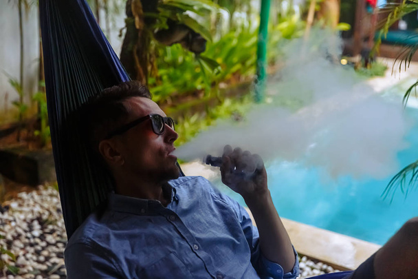Hombre descansando en una hamaca y vapeando y soltando vapor de un cigarrillo electrónico
 - Foto, imagen