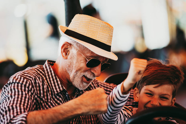 Grandfather and grandson having fun and spending good quality time together in amusement park. They enjoying and smiling while driving bumper car together. - Foto, imagen