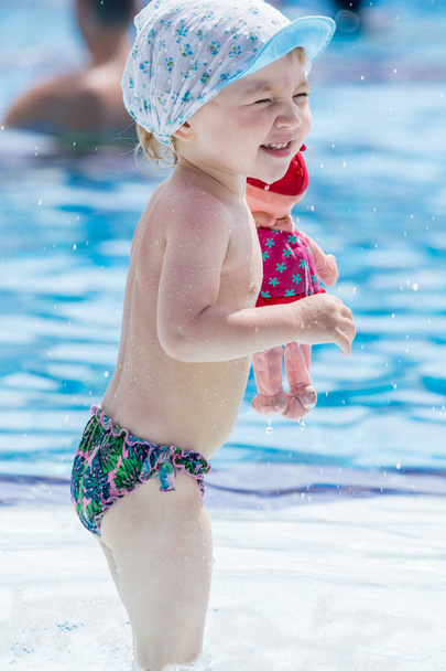 Beautiful child in shorts and cap with doll in his hand squints from sun in blue pool. - Photo, Image