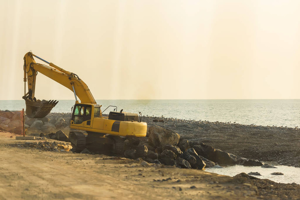 Κίτρινο τρακτέρ που σκάβει στη μαύρη θάλασσα. Περπατήστε κατά μήκος του περιπάτου στο Μπατούμι. Κατασκευαστικές εργασίες κοντά στη θάλασσα. Ανάπαυση στη γεωργία. - Φωτογραφία, εικόνα