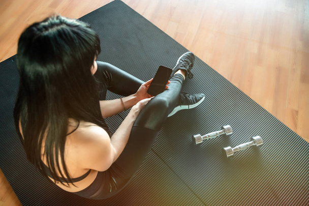 Sporty girl sitting on the floor in the gym with dumbbells and phone. Top view - Photo, image