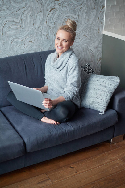 Middle aged woman sitting on sofa in her living room and working on laptop - Photo, Image