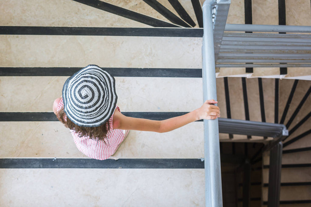 Little girl in striped dress and summer hat comes downstairs holding hand over the railing - Foto, afbeelding