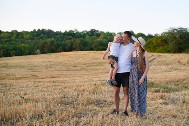 glückliche junge Familie. Ein Vater mit einem kleinen Sohn im Arm und eine schwangere Mutter. abgeschrägtes Weizenfeld im Hintergrund. Sonnenuntergang. - Foto, Bild