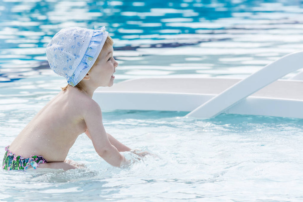 Beautiful child in shorts and hat plays with water in blue pool near white chaise lounge. - Photo, Image
