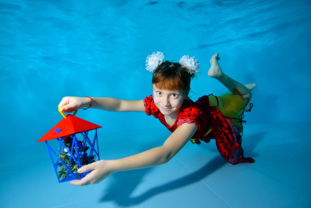 A little girl swims underwater in the pool with her eyes open near the bottom in a red dress, holding a gift in her outstretched hand and smiling. Portrait. Horizontal view - Foto, immagini