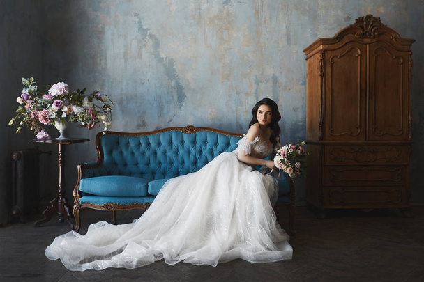 Sexy and beautiful brunette model girl in stylish and fashionable lace wedding dress with naked shoulders sits on the antique sofa and posing in luxury vintage interior - Valokuva, kuva