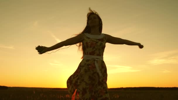 ヘッドフォンの少女と暖かい日没の光の下で踊りながら回転するスマートフォン幸せな女の子は音楽を聞いて、空に対して美しい夕日の光線で踊ります。スローモーション. - 映像、動画