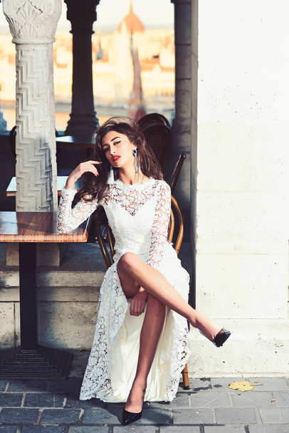 Fashion model with glamour look and makeup. Sexy woman sit alone in street cafe. Woman in white wedding dress outdoor. Girl with long brunette hair. Bride with makeup face - Foto, Imagem