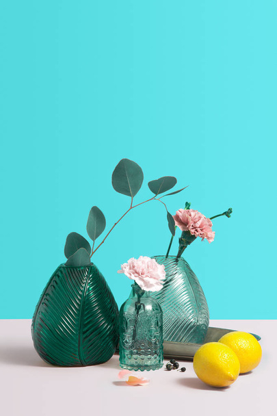 Modern green glass Vase with beautiful pink flowers. Creative Composition with flowers, lemon and vase on blue background. Minimalism with copy space for text or design. Concept for flower shop - Zdjęcie, obraz
