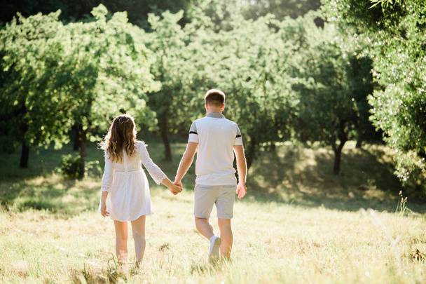 Back view of young pretty couple in love walking  in park. Handsome cheerful blonde girl in white dress hugging her boyfriend. Man and woman having fun outdoors - Φωτογραφία, εικόνα