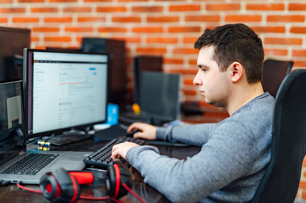 Young smart ambitious programmer using gadgets and working hard in IT company. Programmer is concentrated on problem looking at the screens while sitting in the chair. - Фото, изображение