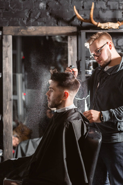 Barber sprays clean water on head in barbershop. Professional trimmer tool cuts beard and hair on young guy in barber shop salon. - Photo, Image