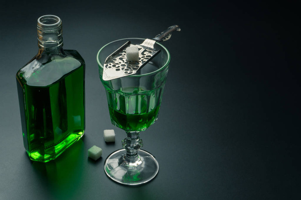 lasillinen absinttia ja ruostumattomasta teräksestä valmistettu ura-lusikka sokerikuutioilla, pöydällä oleva absinttipullo, valikoiva tarkennus
 - Valokuva, kuva
