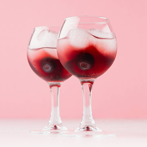 Juicy red arctic traditional cold cocktails with ice cubes, blueberry in wet wineglasses on light pastel pink background, square. - Photo, image