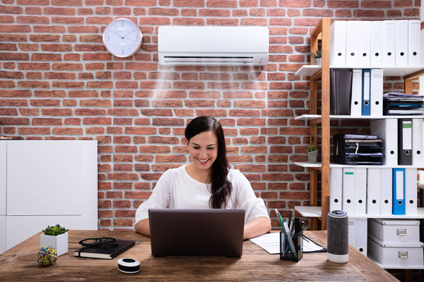 Smiling Businesswoman Enjoying The Cooling Of Air Conditioner Using Laptop At Workplace - Foto, imagen