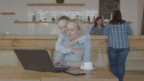 Child hugging mother networking on laptop in cafe - Imágenes, Vídeo