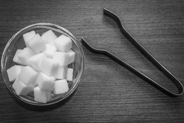 Vista superior de cubos de açúcar em uma tigela de vidro com um par de pinças de cozinha foreceps mantidos no chão de madeira. Configuração de cozinha contrastante preto e branco
. - Foto, Imagem