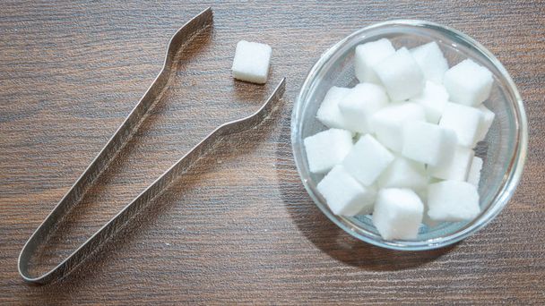 Top view of sugar cubes in a glass bowl with a pair of kitchen tongs foreceps kept on wooden floor. Sugar cube on wooden floor ready to be picked up by tongs - Photo, Image