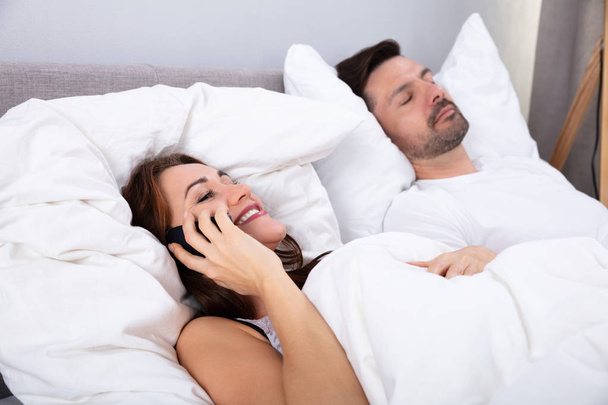 Hymyilevä nuori nainen puhuu älypuhelimella lisäksi hänen miehensä nukkuu sängyssä makuuhuoneessa
 - Valokuva, kuva