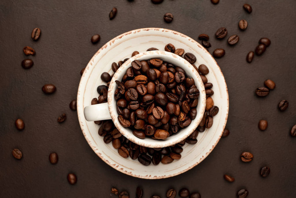 Taza de cerámica y platillo lleno de granos de café tostados sobre fondo de piedra marrón
  - Foto, Imagen