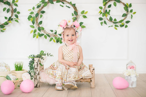 Cute little girl in a dress with a flower print and a hat with flowers in the Easter decorations in the studio. Little girl with easter eggs and flowers in a spring studio - Foto, Bild