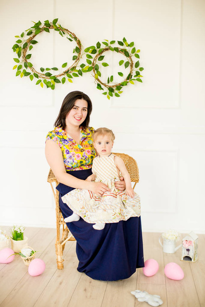 Roztomilá holčička v šatech s květinovým tiskem s mámou v Velikonoční výzdobě ve studiu. Holčička a matka s velikonočními vejci a květinami v jarním studiu - Fotografie, Obrázek