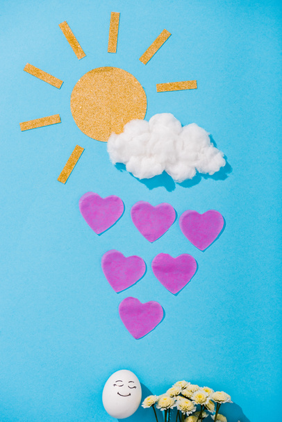 vista superior de sol de papel, nube de algodón de azúcar, huevo con expresión de cara feliz, flores y gotas de lluvia en forma de corazón en azul
 - Foto, Imagen