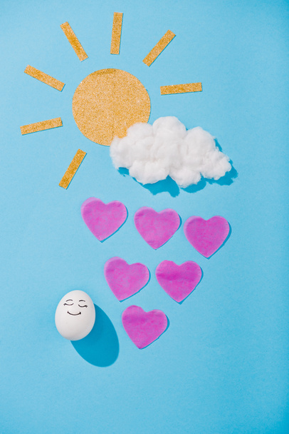 Papiersonne, Zuckerwatte-Wolke, Ei mit glücklichem Gesichtsausdruck und herzförmigen Regentropfen auf Blau - Foto, Bild