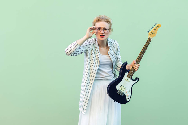 young blonde woman in white clothes and striped blouse with electric guitar in hand holding eyeglasses and looking attentive at camera on light green background. - Foto, Bild