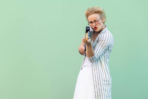 young blonde woman in white clothes and striped blouse with eyeglasses standing holding electric guitar like gun and focusing on camera over light green background. - Zdjęcie, obraz