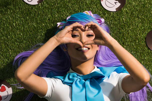 Sonriendo chica de anime asiático acostado en la hierba y mostrando signo de corazón
 - Foto, Imagen