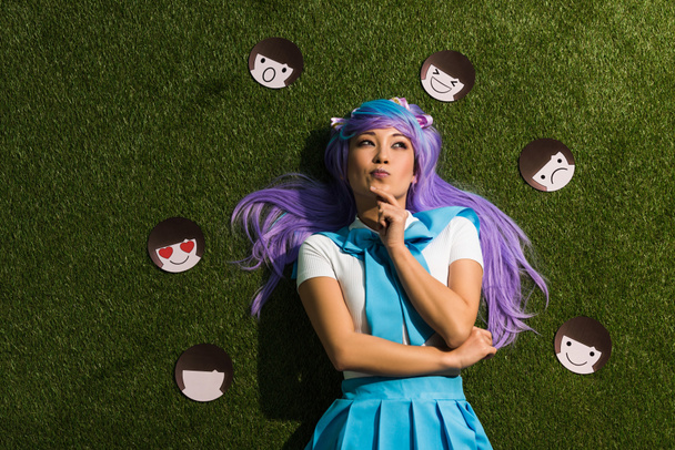Pensivo asiático anime menina em roxo peruca com emoticons deitado na grama
 - Foto, Imagem