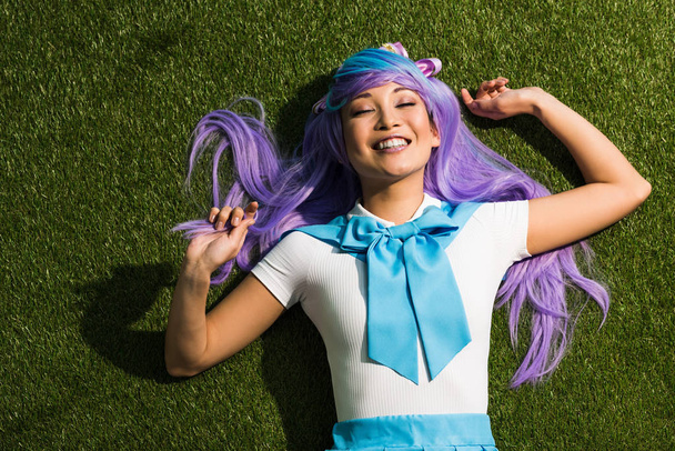 Laughing anime girl in purple wig lying on grass with closed eyes - Photo, Image