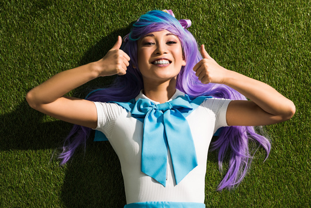 Lachende Aziatische anime meisje liggend op gras en toont duimen omhoog - Foto, afbeelding