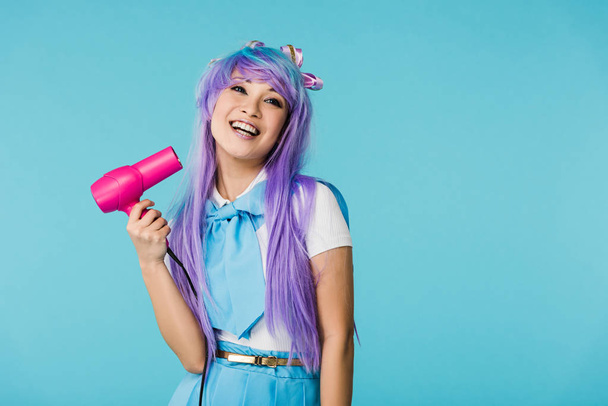 Smiling asian anime girl in wig using hairdryer isolated on blue - 写真・画像