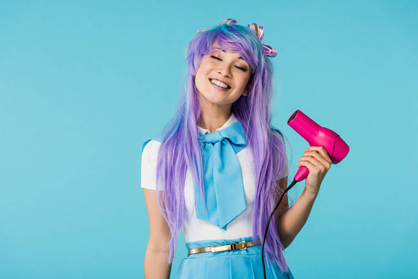 Sorrindo anime menina na peruca usando secador de cabelo com olhos fechados isolado em azul
 - Foto, Imagem