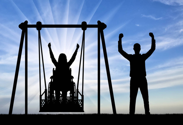Sylwetka szczęśliwej pary, kobieta na wózku inwalidzkim na adaptacyjnej huśtawce i zdrowego człowieka w pobliżu. - Zdjęcie, obraz