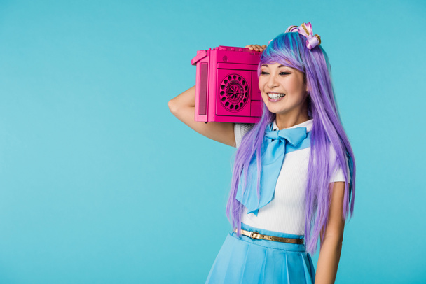 Улыбающаяся азиатская девушка отаку в фиолетовом парике держит бумбокс изолированный на голубом
 - Фото, изображение