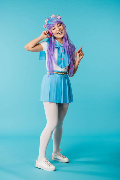 Full length view of anime girl in wig listening music in headphones with closed eyes on blue - Zdjęcie, obraz