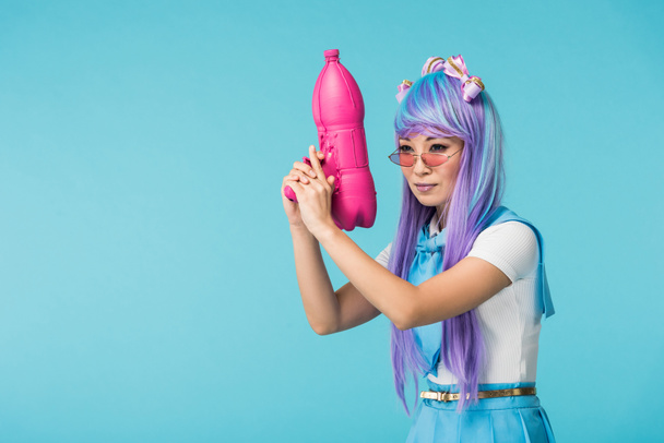 Asian anime girl in wig and glasses holding water gun isolated on blue - Fotó, kép