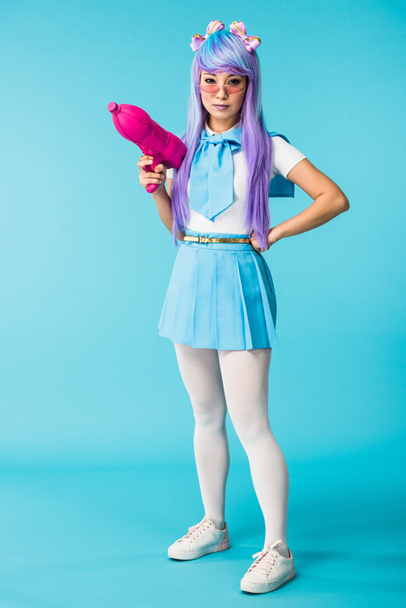 Visão de comprimento total da menina anime asiática em peruca e óculos segurando arma de água no azul
 - Foto, Imagem